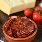 closeup of an earthenware bowl with some slices of Spanish chorizo, a pork sausage typical of Spain, on a rustic wooden table with a piece of cheese, some tomatoes and garlic cloves in the background